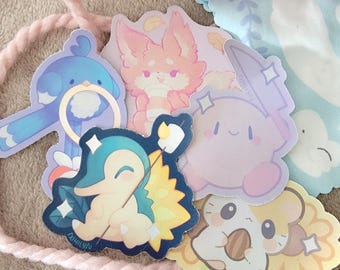 Puede incluir: Una colección de pegatinas coloridas con animales y personajes de dibujos animados. Las pegatinas incluyen un pájaro azul, un zorro rosa, un personaje lila claro, un hámster y una criatura azul y amarilla. Las pegatinas están sobre una cuerda rosa claro.