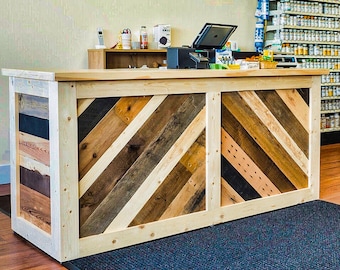 May include: A wooden counter with a chevron pattern made from reclaimed wood. The counter is painted white and has a natural wood top. The counter is in a store with shelves of products in the background.
