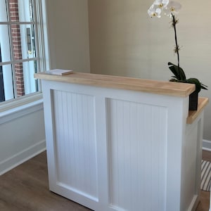 May include: A white reception desk with a light wood top and vertical paneling. A white orchid in a black pot sits on the right side. A window is visible in the background.