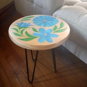 May include: A round wooden side table with a floral design. The tabletop features hand-painted blue flowers and green leaves on a light wood surface. The table has three black hairpin legs.