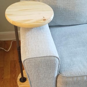 May include: A side table featuring a circular light-colored wood tabletop and a matching circular wood base connected by a dark metal pipe support. The table is designed to slide over the arm of a sofa, providing a flat surface for items.