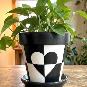 May include: A black and white checkered plant pot with a solid black rim and a matching black saucer. The exterior features a large heart shape integrated into the checkerboard pattern. A green leafy plant grows from the top of the container.