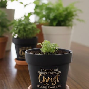 May include: A black plant pot with a small green plant inside, sitting on a terracotta saucer. The pot has gold lettering that reads "I can do all things through Christ who strengthens me." Other potted plants are in the background.