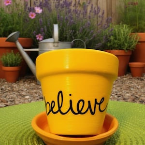 May include: A bright yellow painted flower pot with the word "believe" in black script. The pot sits on a matching orange saucer. The background includes other terracotta pots, plants, and a watering can.