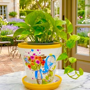 May include: A white ceramic plant pot with a bright yellow rim and matching saucer, featuring a colorful painted graphic of a blue bicycle carrying a basket of vibrant flowers. A lush green pothos plant grows from the top of the container.