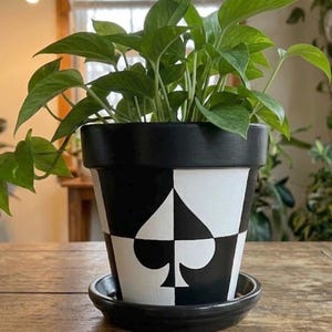 May include: A black and white terracotta plant pot featuring a spade symbol from a deck of cards. The pot has a solid black rim and base, with a geometric checkerboard pattern around the centre. A green pothos plant grows from the top.
