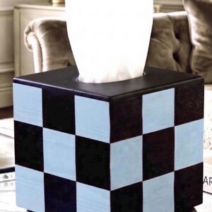 May include: A square tissue box cover featuring a black and light blue checkered pattern on all visible sides. The box is supported by four small, round, black feet at the base and has a central opening at the top with white tissues protruding.