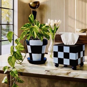 May include: A square tissue box cover featuring a black and light blue checkerboard pattern, supported by four small spherical feet. The box holds white tissues and is displayed alongside a matching black and light blue checkered plant pot.