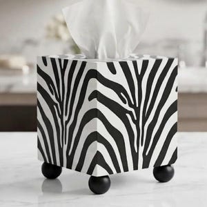 May include: A square tissue box cover featuring a black and white zebra stripe pattern. The box has a flat top with an opening for tissues and is supported by four small, round, black feet at the base. A white tissue is pulled through the top.