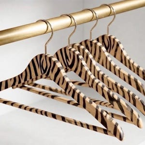 May include: A set of four wooden clothes hangers featuring a black and tan zebra stripe pattern. Each hanger has a curved top, a horizontal bottom bar, and a metallic gold-toned hook, all suspended from a matching gold-toned closet rod.