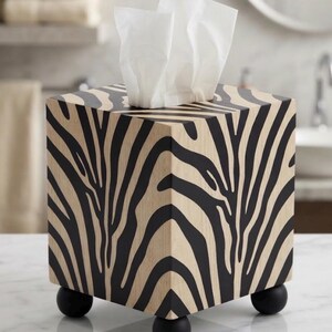 May include: A square tissue box cover featuring a black and beige zebra stripe pattern on all visible sides. The box is elevated by four small, round, matte black feet and contains a stack of white facial tissues protruding from the top opening.
