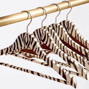 May include: A set of four clothes hangers featuring a black and white zebra stripe pattern. Each hanger is constructed with a curved shoulder frame, a horizontal trouser bar, and a silver-toned metal hook, all suspended from a gold-toned rod.