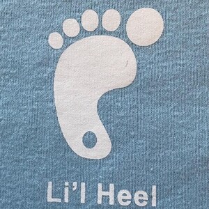 Puede incluir: Una camiseta azul claro con un gr&aacute;fico de huella de pie blanco y el texto "Lil' Heel" impreso en la parte delantera.