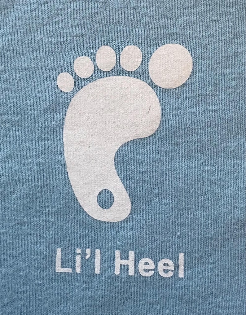 Puede incluir: Una camiseta azul claro con un gr&aacute;fico de huella de pie blanco y el texto "Lil' Heel" impreso en la parte delantera.