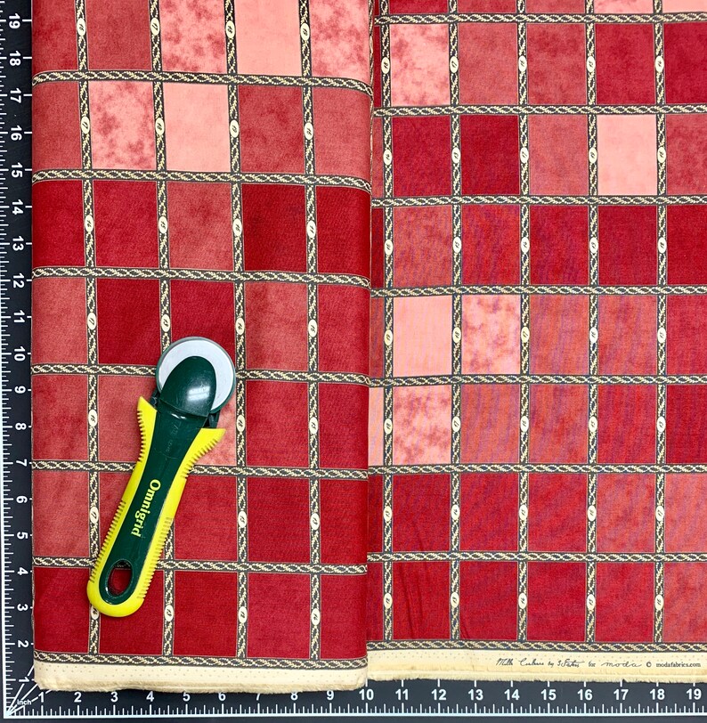 Puede incluir: Tela roja y rosa con un patr&oacute;n a cuadros. La tela tiene un borde de dise&ntilde;o de cuerda. Un cortador rotatorio est&aacute; encima de la tela.