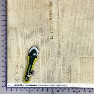 Pode incluir: Um tecido bege com uma colagem impressa de documentos hist&oacute;ricos com texto incluindo "IN CONGRESS" e "We the People". Um cortador rotativo amarelo e preto est&aacute; colocado sobre o tecido ao lado de uma r&eacute;gua que mostra 43 cent&iacute;metros.