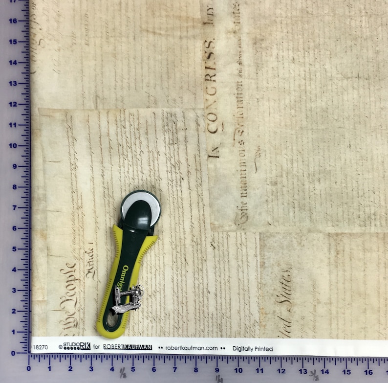 Pode incluir: Um tecido bege com uma colagem impressa de documentos hist&oacute;ricos com texto incluindo "IN CONGRESS" e "We the People". Um cortador rotativo amarelo e preto est&aacute; colocado sobre o tecido ao lado de uma r&eacute;gua que mostra 43 cent&iacute;metros.