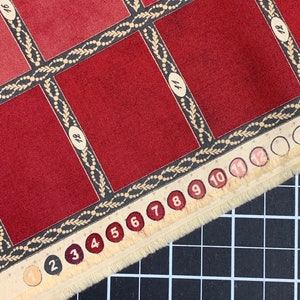Puede incluir: Una muestra de tela roja con un patr&oacute;n de cuadr&iacute;cula y los n&uacute;meros del 1 al 12 impresos en el borde. La tela tiene un borde decorativo con un dise&ntilde;o de hojas y cuentas.