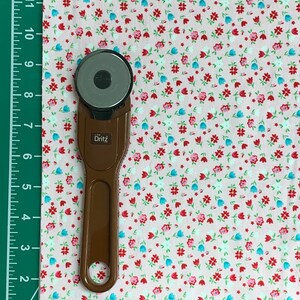 May include: A brown rotary cutter with a silver blade on a pink fabric with a small white, red, and blue floral pattern. The fabric is measured with a ruler in inches.