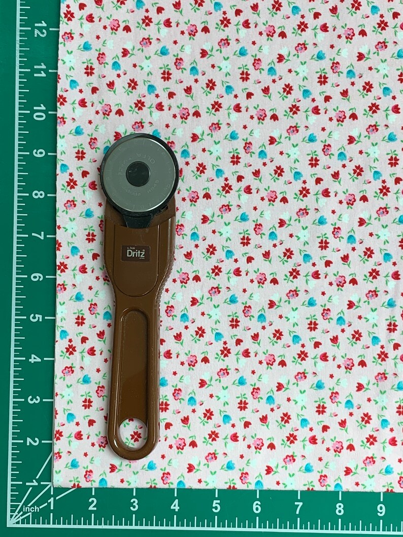 May include: A brown rotary cutter with a silver blade on a pink fabric with a small white, red, and blue floral pattern. The fabric is measured with a ruler in inches.
