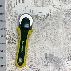 May include: A green and yellow rotary cutter with a black handle and the word "Omnigrid" printed on the side. The cutter is on top of a white fabric with a newspaper print pattern.