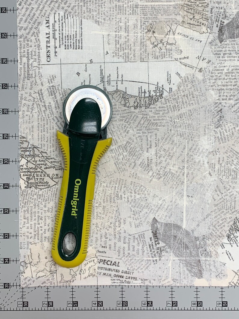 May include: A green and yellow rotary cutter with a black handle and the word "Omnigrid" printed on the side. The cutter is on top of a white fabric with a newspaper print pattern.