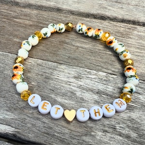 Può includere: Un braccialetto di perline bianche con un motivo floreale e dettagli dorati. Il braccialetto recita "LET THEM" in lettere dorate, con un ciondolo a forma di cuore dorato. Le perline floreali hanno fiori gialli e arancioni.
