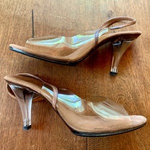 May include: A pair of clear plastic heeled sandals with a brown suede insole and a slingback strap. The heels are clear plastic and have a black base.