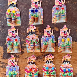 May include: A collection of colorful, handmade paper dolls, each featuring a unique design. The dolls have a floral theme, with small potted flowers and vibrant patterns. The dolls are arranged on a wooden surface.