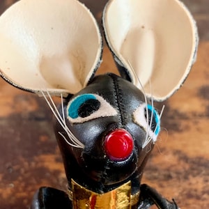 May include: A handcrafted mouse figurine with large, cream-coloured ears and a black body. The mouse has blue eyes, a red nose, and a gold bow tie. White whiskers extend from the face.