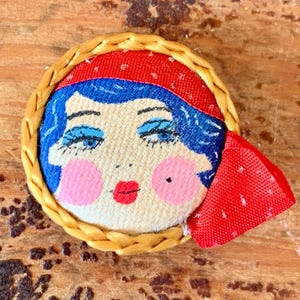 May include: A circular brooch featuring a stylized illustration of a face with blue hair, a red headband, and bright pink cheeks. The face is framed by a braided tan border, and a red fabric accent is attached to the right side of the piece.