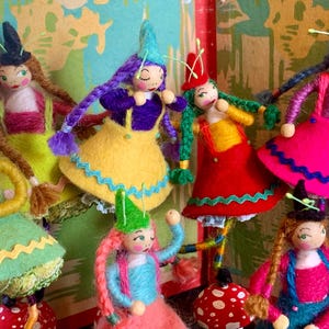 May include: A collection of handmade dolls with whimsical designs. Each doll features a unique outfit in vibrant colors like yellow, pink, and green, with detailed facial features and yarn hair. The dolls have a mushroom-shaped base and are adorned with small hats.