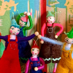 May include: A group of whimsical handmade dolls with painted faces, wearing colorful felt overalls and hats. The dolls have arms outstretched and are posed in a playful arrangement. The color palette includes red, green, blue, and yellow.