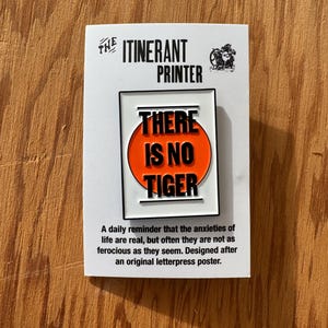 May include: A white enamel pin with a red circle and black text that reads "THERE IS NO TIGER". The pin is on a white card with black text that reads "THE ITINERANT PRINTER" and a description of the pin.