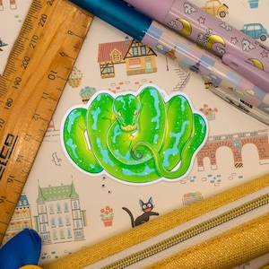 May include: A green and blue snake sticker with a sparkly texture. The sticker is on a light brown background with a pattern of houses, cats, and flowers.