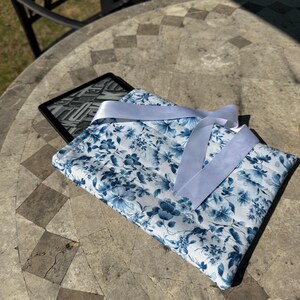Könnte beinhalten: Eine Kindle-Hülle mit weißem und blauem Blumenmuster und passendem Bandverschluss. Die Hülle ist gesteppt und hat ein Muster aus blauen Blumen und Blättern. Ein E-Reader ist im Inneren zu sehen.