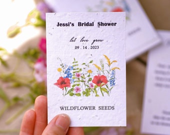May include: White paper card with wildflower seeds. The card has a watercolor illustration of wildflowers and the text "Jessi's Bridal Shower let love grow 09.14.2023 WILD FLOWER SEEDS".