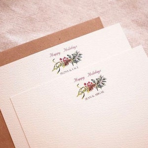 May include: Two white cards with a brown envelope. The cards have a watercolor illustration of a sprig of holly with red berries and the text "Happy Holidays" in pink. The top card has the text "Penny & Lace" and the bottom card has the text "Sean & Abigail".