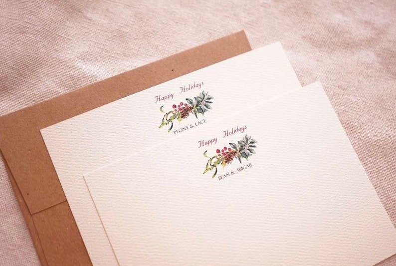 May include: Two white cards with a brown envelope. The cards have a watercolor illustration of a sprig of holly with red berries and the text "Happy Holidays" in pink. The top card has the text "Penny & Lace" and the bottom card has the text "Sean & Abigail".