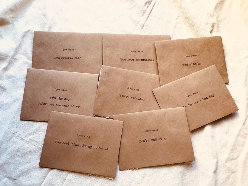 May include: A set of ten brown paper envelopes with handwritten messages on each. The messages include "Open when you receive this", "Open when you're stressed", "Open when you're having a bad day", "Open when you miss me", "Open when you feel like giving up on us", "Open when you're mad at me", "Open when it's the day before we see each other", and "Open when you need reassurance".