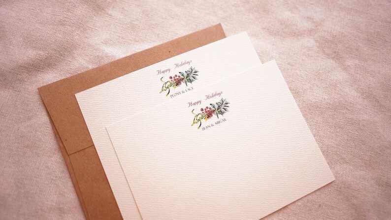 May include: Two white cards with a brown envelope. The cards have a floral design with red berries and green leaves. The text on the cards reads "Happy Holidays".
