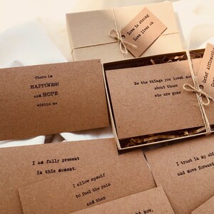 Puede incluir: Un juego de tarjetas de afirmaci&oacute;n marrones con mensajes positivos impresos en ellas. Las tarjetas est&aacute;n empaquetadas en una caja marr&oacute;n con una cuerda marr&oacute;n y una etiqueta que dice "Conjunto de tarjetas de afirmaci&oacute;n de duelo".