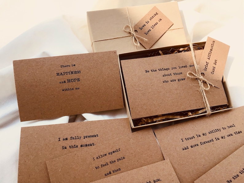 Puede incluir: Un juego de tarjetas de afirmaci&oacute;n marrones con mensajes positivos impresos en ellas. Las tarjetas est&aacute;n empaquetadas en una caja marr&oacute;n con una cuerda marr&oacute;n y una etiqueta que dice "Conjunto de tarjetas de afirmaci&oacute;n de duelo".