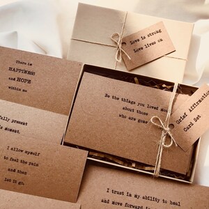 Puede incluir: Un conjunto de tarjetas de papel marr&oacute;n con afirmaciones positivas para el duelo. Las tarjetas est&aacute;n atadas con un cordel y vienen en una caja marr&oacute;n con una etiqueta de papel marr&oacute;n que dice "Conjunto de tarjetas de afirmaci&oacute;n del duelo".