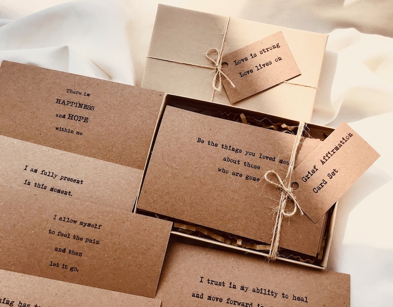 Puede incluir: Un conjunto de tarjetas de papel marr&oacute;n con afirmaciones positivas para el duelo. Las tarjetas est&aacute;n atadas con un cordel y vienen en una caja marr&oacute;n con una etiqueta de papel marr&oacute;n que dice "Conjunto de tarjetas de afirmaci&oacute;n del duelo".