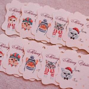 May include: A collection of cream-colored gift tags with scalloped edges, featuring watercolor illustrations of cats in winter hats and scarves. The tags read "Meowy Christmas" and have "From:" and "To:" fields.