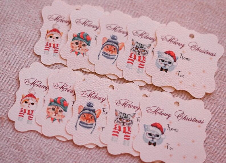 May include: A collection of cream-colored gift tags with scalloped edges, featuring watercolor illustrations of cats in winter hats and scarves. The tags read "Meowy Christmas" and have "From:" and "To:" fields.