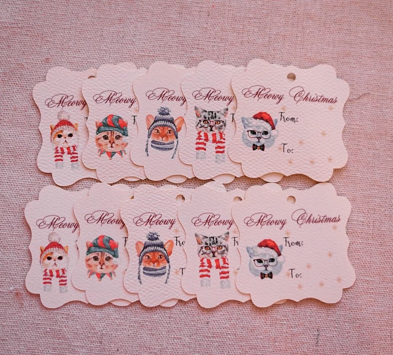 May include: A collection of cream-colored gift tags with scalloped edges. Each tag features a watercolor illustration of a cat wearing a hat and scarf. The tags are labeled "Meowy Christmas" with "From:" and "To:" fields.