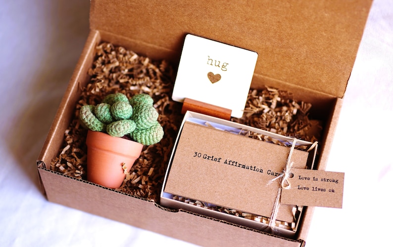 Puede incluir: Una caja de cart&oacute;n marr&oacute;n llena de papel de embalaje marr&oacute;n. La caja contiene una peque&ntilde;a planta en maceta, una tarjeta blanca con la palabra "hug" y un coraz&oacute;n, y una caja de 30 tarjetas de afirmaci&oacute;n de duelo con una etiqueta que dice "El amor es fuerte, El amor sigue vivo".