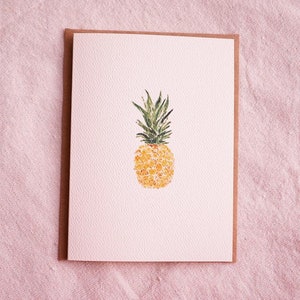 May include: A watercolor painting of a pineapple on a white card with a brown border. The pineapple is yellow with green leaves.